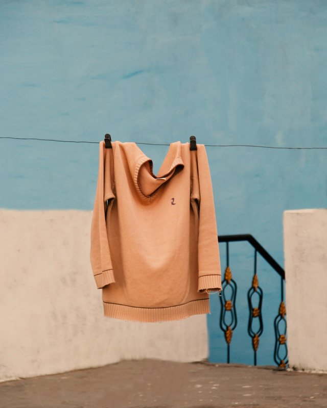 A tan knitted hoodie hanging from a clothesline, the hood folded over the line, against a painted wall.